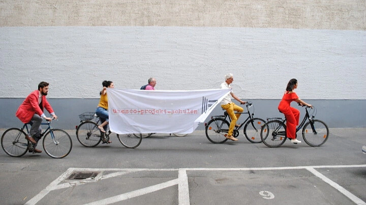 Eine Gruppe von 5 Personen fahren Fahrrad und die in der Mitte halten ein Banner noch auf dem steht: UNESCO-Projekt-Schulen